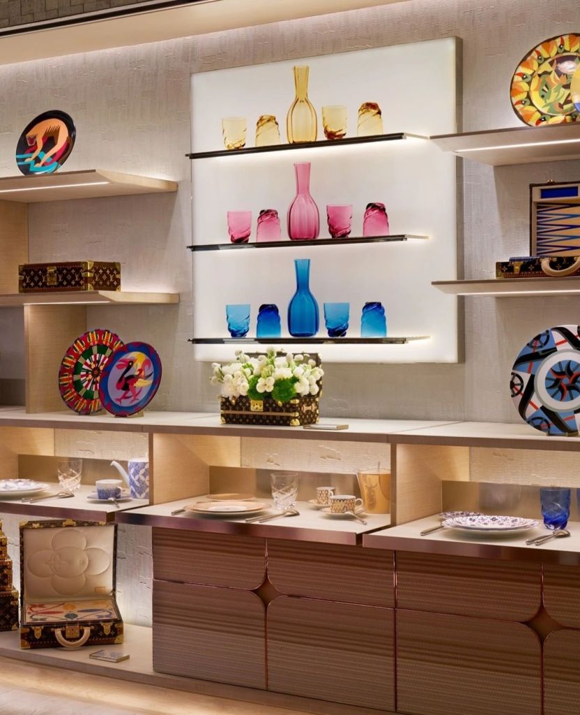 A display of colorful glassware, including vases and glasses, arranged on shelves, alongside decorative plates and floral arrangements, set in a modern interior.