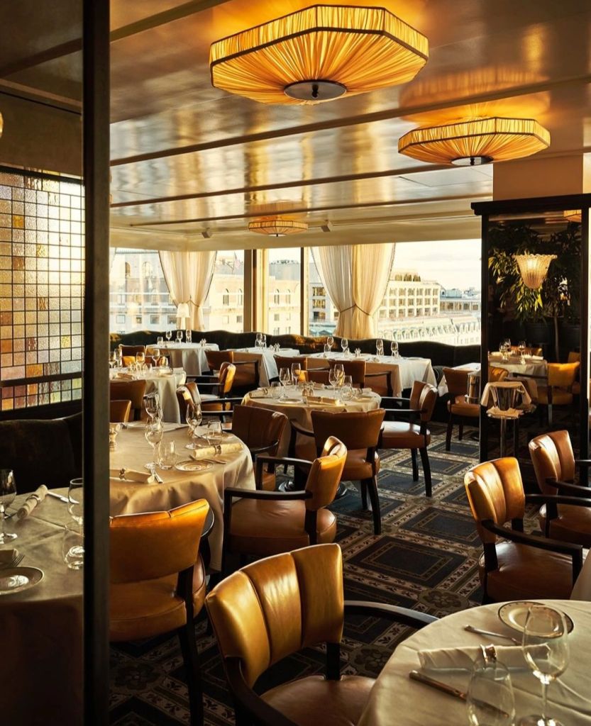 Interior view of a luxurious dining room featuring elegantly set tables, plush leather chairs, and stylish light fixtures, with large windows showcasing a scenic view.