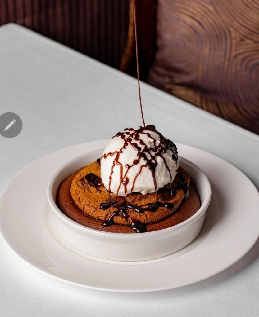 A delicious dessert featuring a warm chocolate chip cookie topped with a scoop of vanilla ice cream and drizzled with chocolate sauce, served in a white bowl on a round plate.