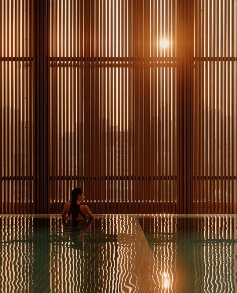 A serene sunset view of a woman sitting by the edge of an infinity pool, surrounded by warm wooden slats that create a peaceful and tranquil atmosphere.
