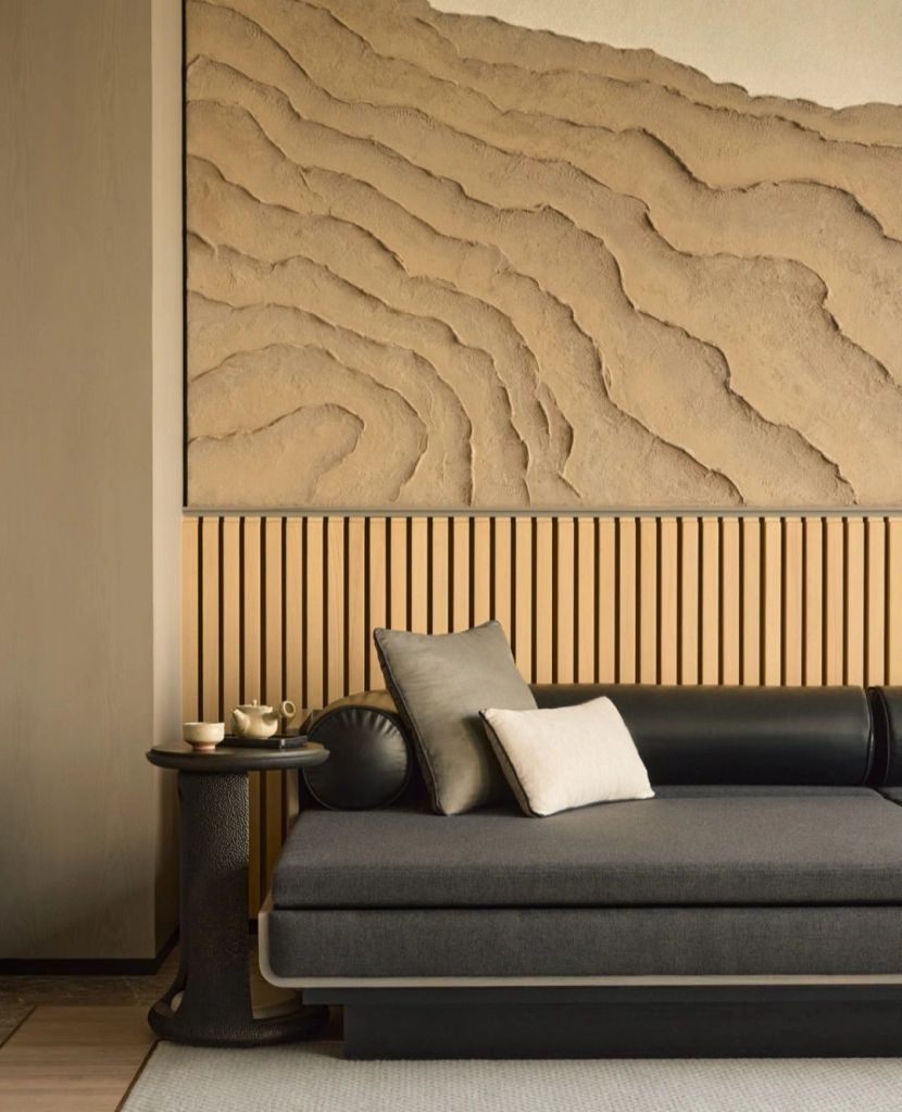 A cozy seating area featuring a dark gray sofa adorned with various cushions, next to a small round table with delicate tea set, and a textured wall art depicting soft, wavy patterns.