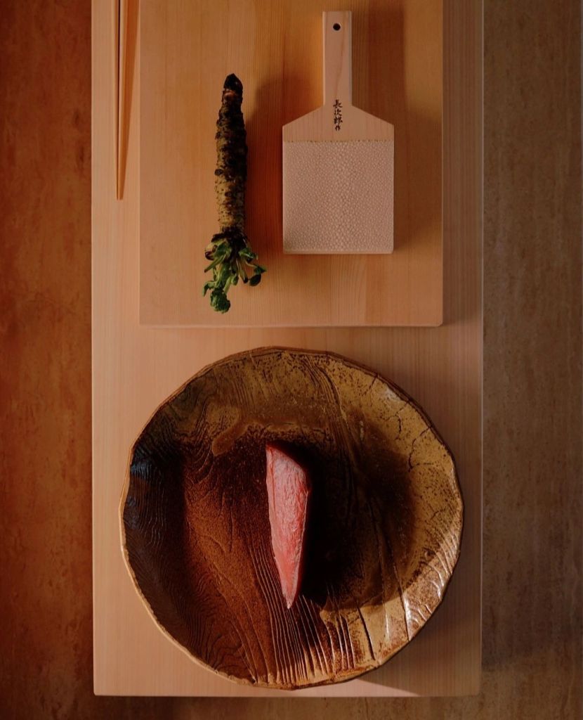 An artistic presentation of a slice of sashimi on a textured brown plate, accompanied by a piece of fresh wasabi and a traditional grater, set on a light wooden surface.