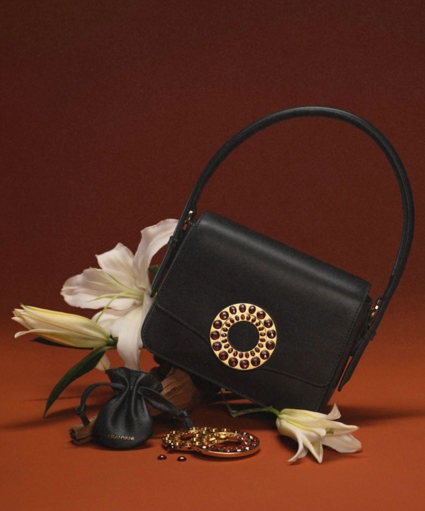 A luxurious black handbag with a gold circular embellishment adorned with red jewels, placed beside white lilies on a warm, earthy background.