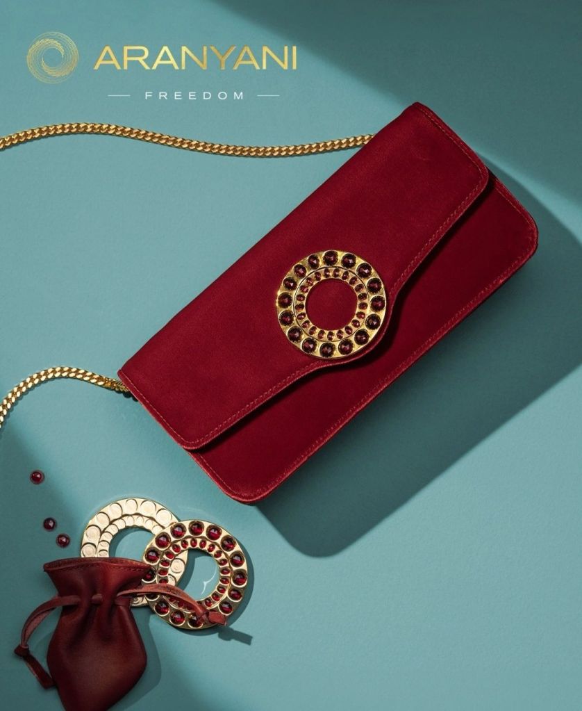 A luxurious red handbag from Aranyani featuring a decorative gold medallion adorned with gemstones, accompanied by two circular accessories and set against a soft blue background.