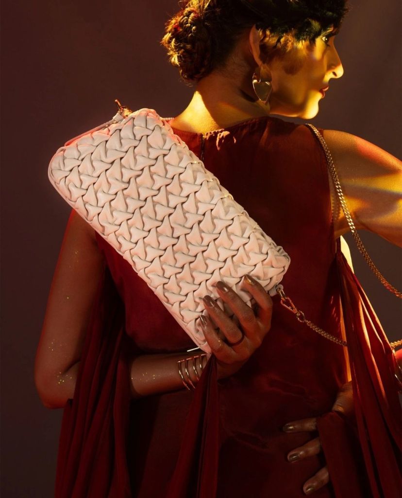 A model elegantly poses in a stylish red ensemble, showcasing a luxurious white handbag with intricate woven detailing. The background features warm lighting that accentuates the sophistication of the look.