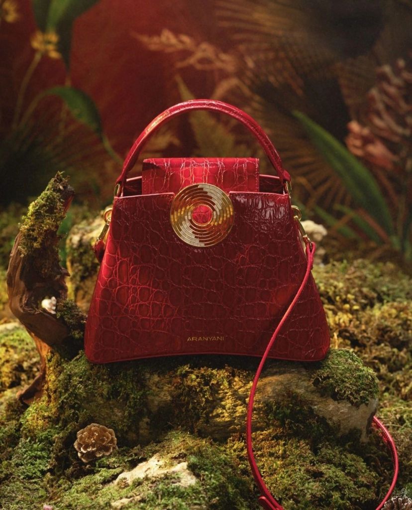 A luxurious red handbag from Aranyani Atelier, featuring a unique design with a circular gold embellishment, placed on a natural mossy background.