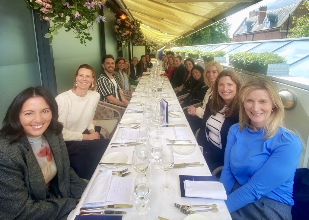 A group of professionals gathered around a beautifully set dining table, sharing smiles and conversation in an elegant outdoor setting, surrounded by greenery and flowers.