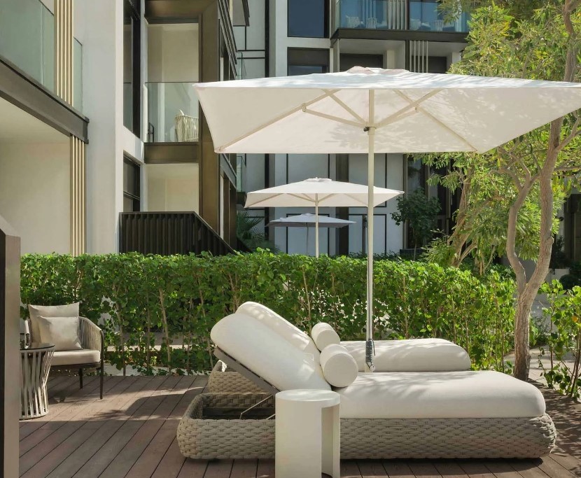 A luxurious outdoor lounge area with a white sun lounger, a small white table, and white umbrellas, surrounded by greenery and modern architecture.