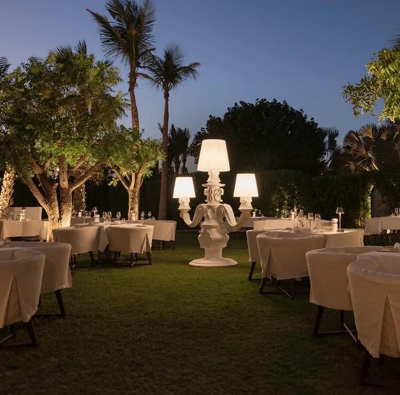 An outdoor dining area in Delano Dubai featuring elegantly set tables under palm trees, illuminated by a unique white chandelier with large lamp shades at dusk.