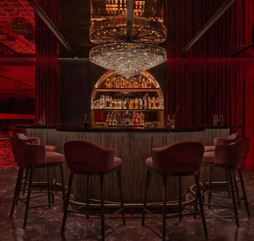 Chic bar interior featuring a large crystal chandelier, plush red seating, and a well-stocked liquor shelf against a backdrop of rich red curtains.