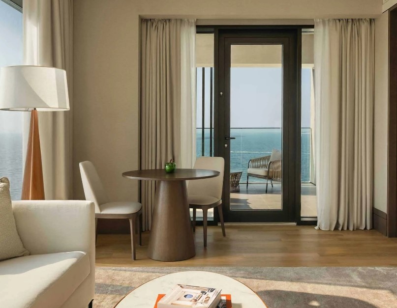 View of a modern hotel room at Delano Dubai, featuring a round table, two chairs, and large glass doors leading to a balcony with an ocean view.