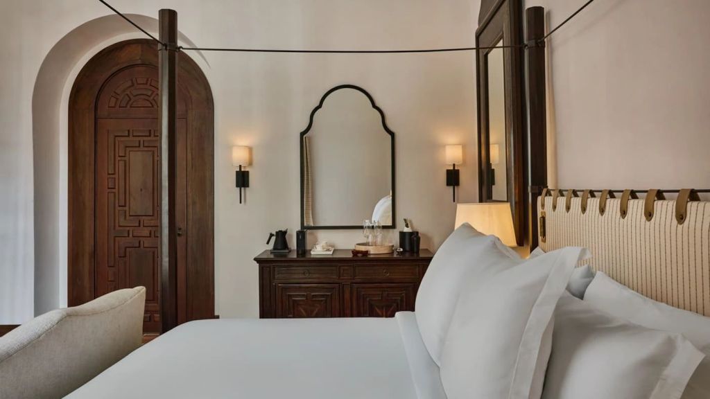 A luxurious hotel room featuring a four-poster bed, elegant furnishings, a wooden door, and ambient lighting, reflecting a blend of modern comfort and heritage style.