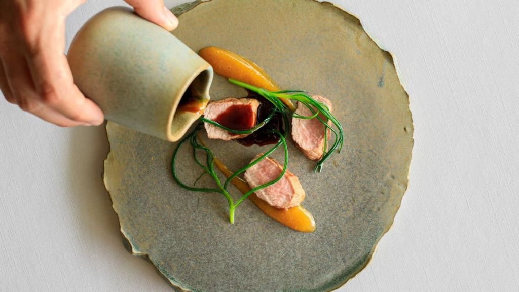 A chef's hand pouring sauce over a beautifully plated dish featuring sliced meat, green onions, and vibrant sauces on a textured ceramic plate.