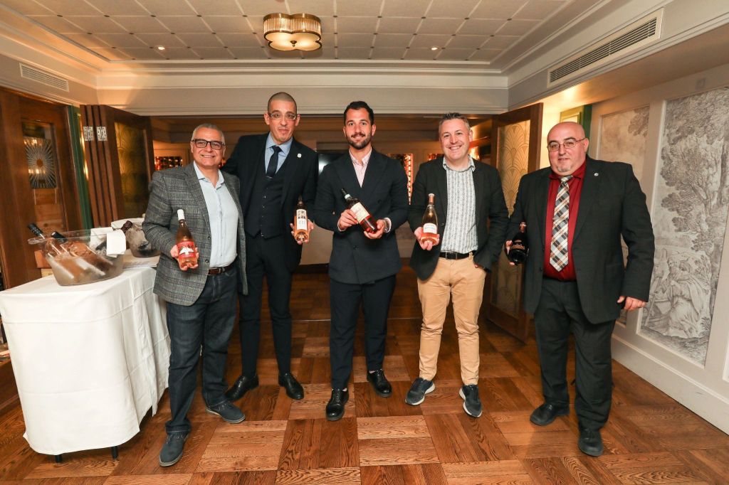 A group of five men stand together in an elegant restaurant setting, holding various bottles of wine. The atmosphere is sophisticated, with wooden flooring and stylish decor in the background.