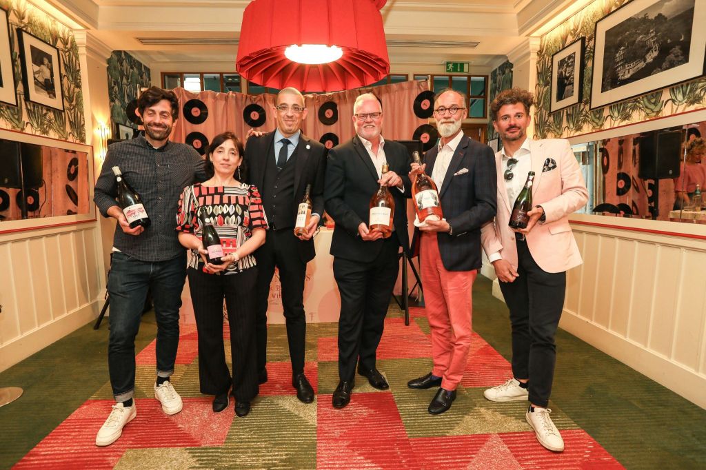 Group of six individuals posing together, holding bottles of wine, in a decorated indoor setting with a red lamp and vinyl records on the wall.