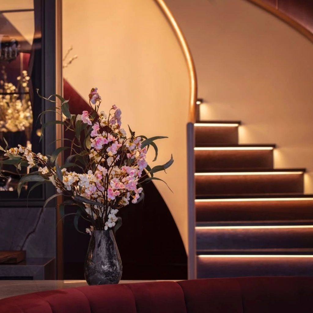 A decorative vase with pink and white flowers sits on a countertop, alongside a plush red seating area, with softly lit stairs in the background.