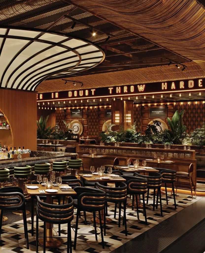 Interior view of Kapitol, a new dining and entertainment venue, showcasing elegant wooden furnishings, stylish seating, and lush greenery under ambient lighting.