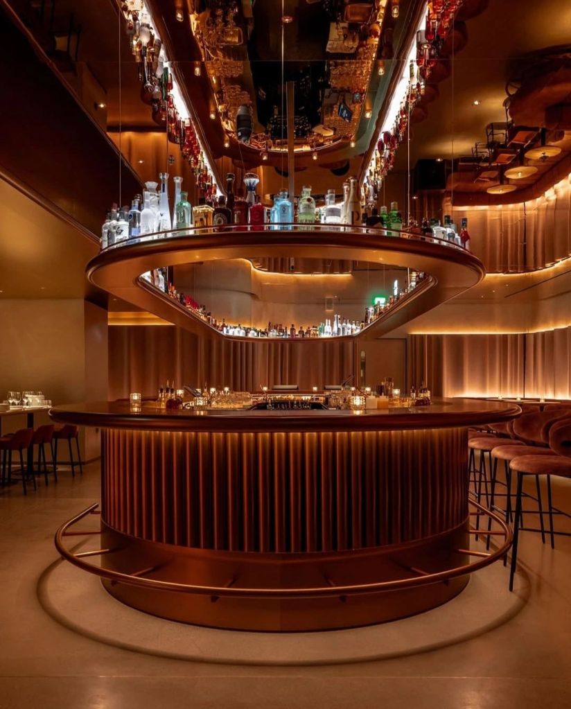 Interior view of a modern bar with a circular design, illuminated by warm lighting and featuring an array of bottles displayed on a mirrored shelf above.