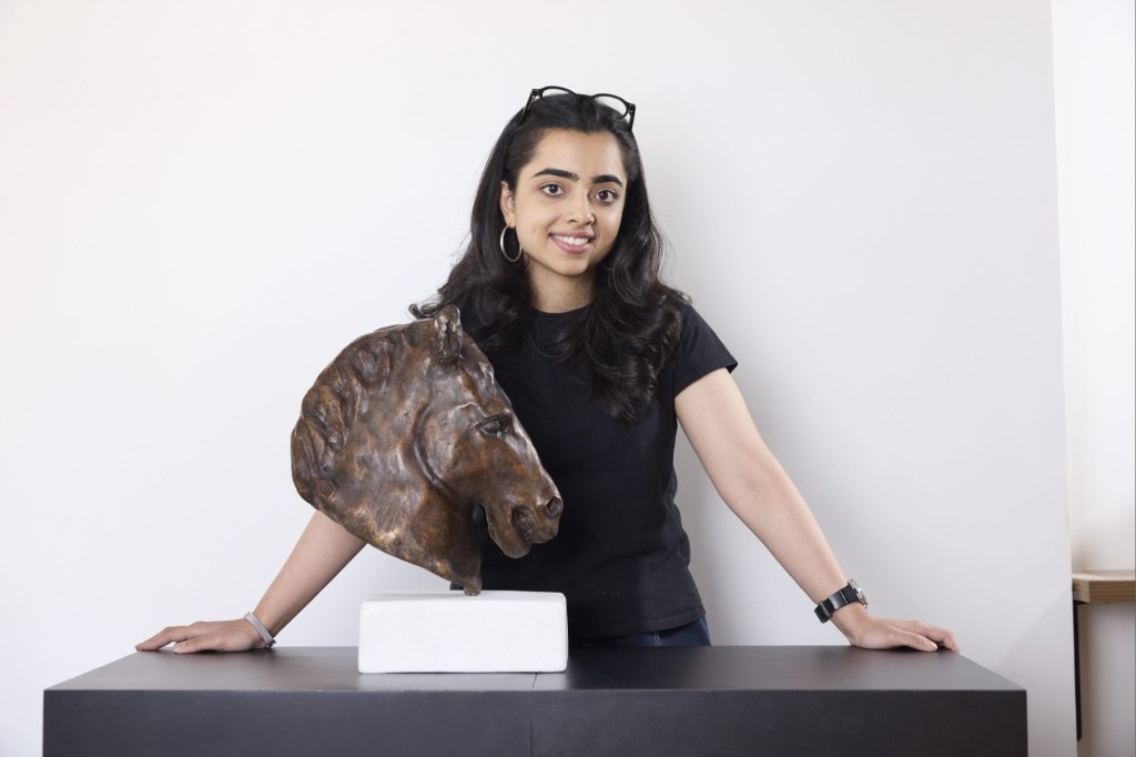 A young woman with long dark hair poses with a bronze sculpture of a horse's head on a pedestal, wearing a black shirt in a bright, minimalistic setting.