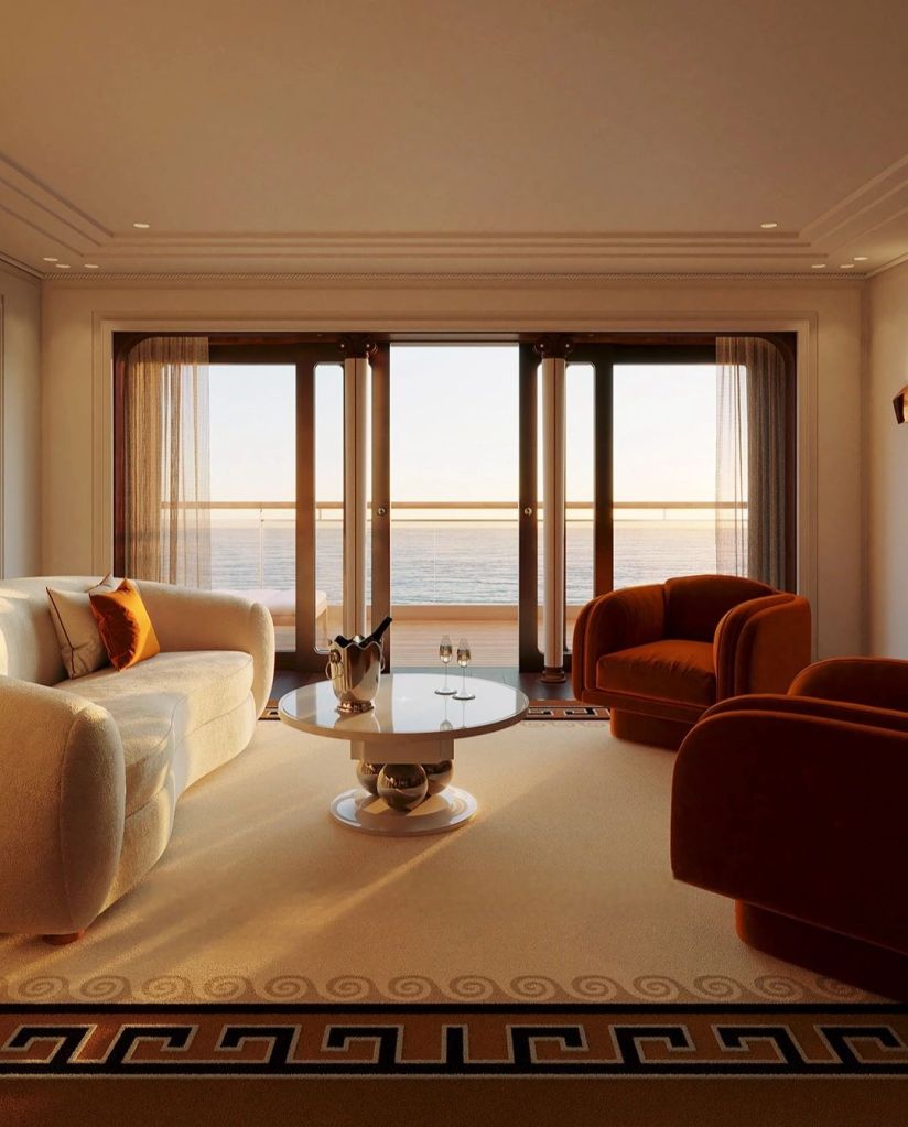 A luxurious yacht suite featuring a plush cream sofa, two orange armchairs, and a round glass table overlooking the sea through large windows, illuminated by warm sunset light.