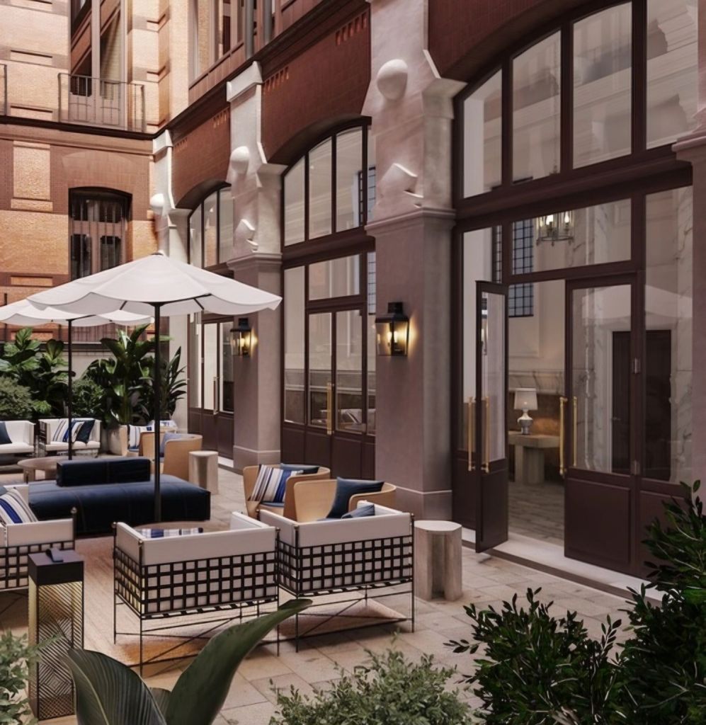 Ground-level view of a contemporary courtyard with modern seating arrangements featuring cushions and a large umbrella, surrounded by lush green plants and large windows of a historic building.