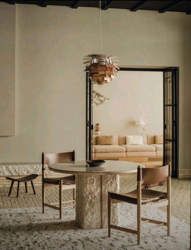 A minimalist dining area featuring a round table with a stone base, two elegant chairs with leather seating, and a unique pendant light fixture above, with a cozy living room visible in the background.