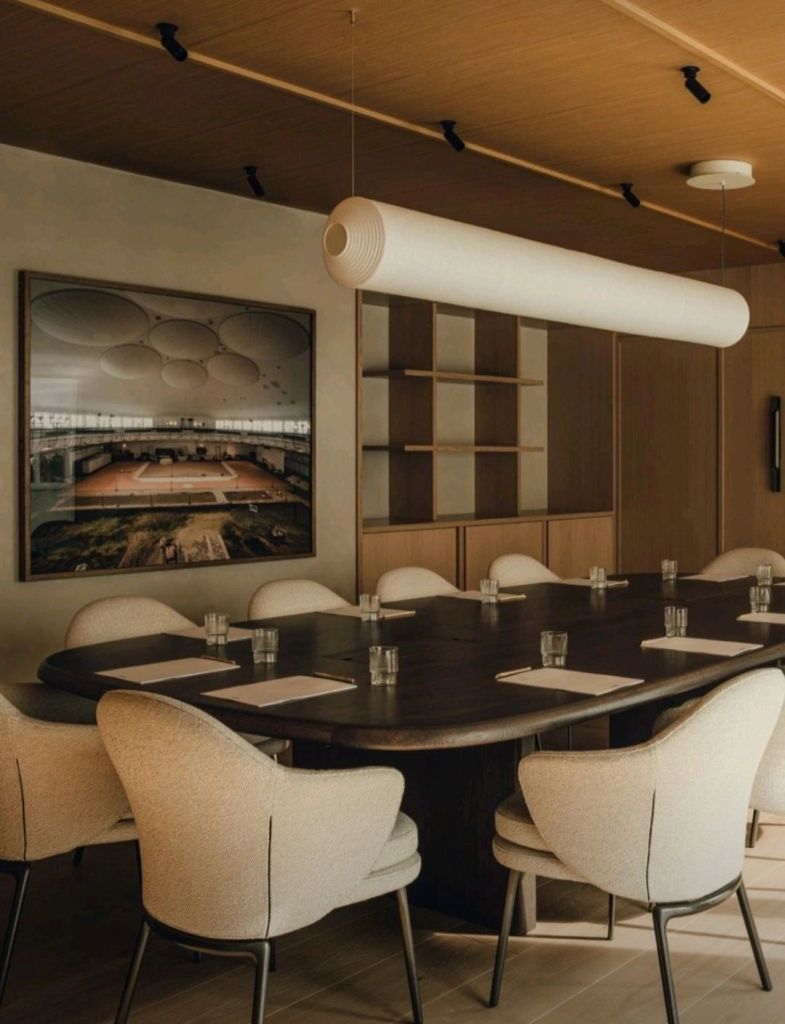 A modern meeting room featuring a large dark wooden table surrounded by light upholstered chairs, illuminated by a stylish hanging light fixture. A large photograph decorates the wall, adding a contemporary touch to the minimalist decor.