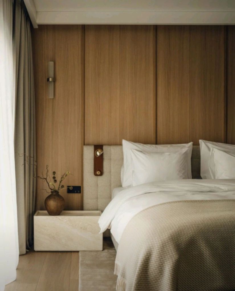 A serene hotel bedroom featuring a plush bed with white linens, a minimalist design, and warm wooden paneling. A bedside table with a decorative vase adds a touch of nature to the space.