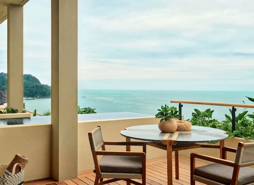 A serene balcony view at Rosewood Mandarina, featuring a round marble table with wooden chairs and potted plants, overlooking the calm ocean and lush greenery.