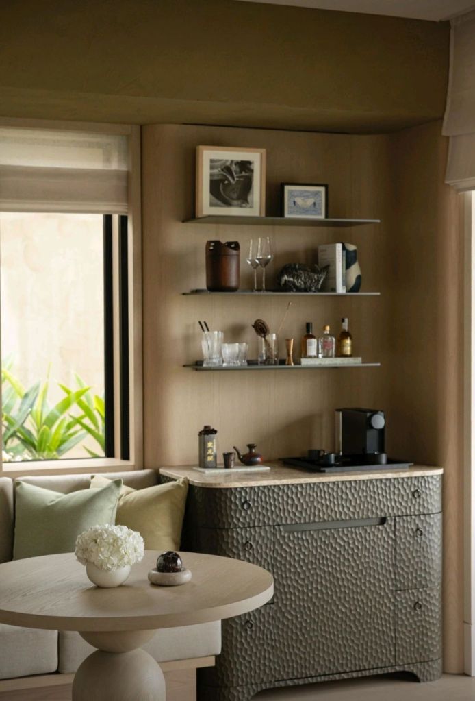 An elegantly designed corner of a luxury hotel room featuring a small round table, a plush sofa with green cushions, and a textured cabinet displaying glassware and decorative items.