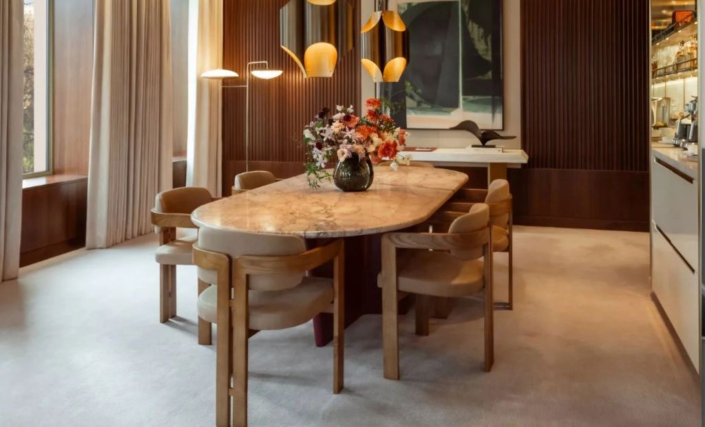 A elegantly designed dining area featuring a large marble table surrounded by modern chairs, with a floral arrangement as a centerpiece and warm lighting fixtures above.