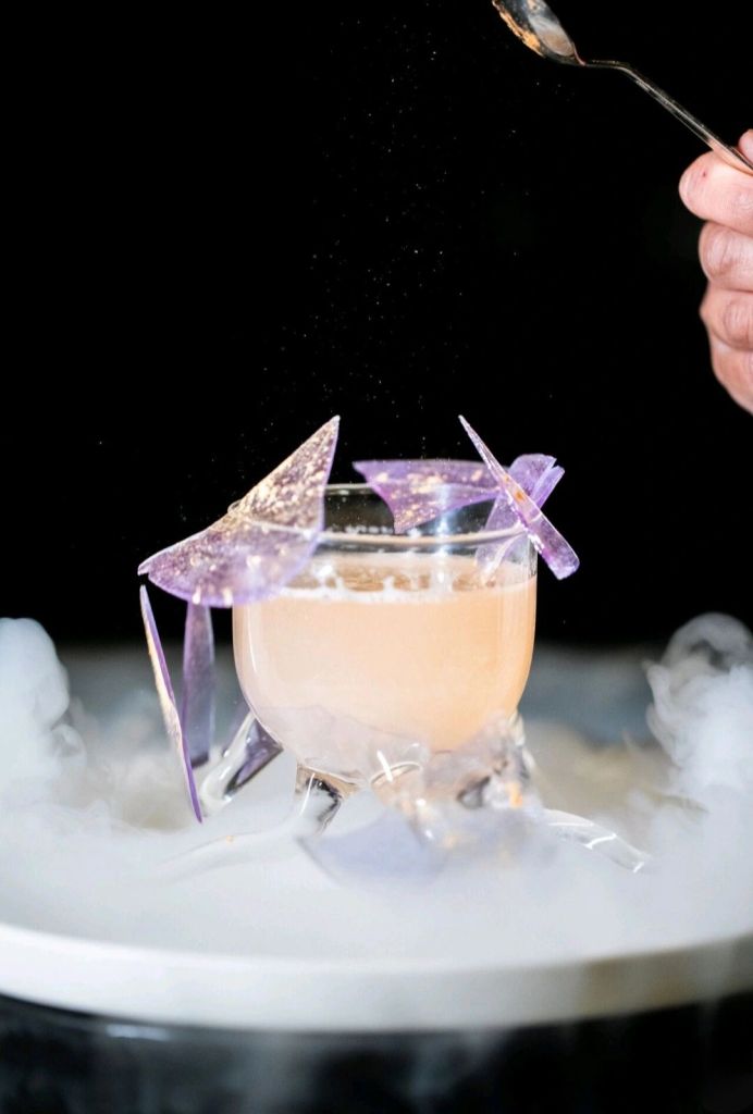 A creative cocktail garnished with colorful, translucent abstract shapes, resting on a frosted surface with vapor effects.