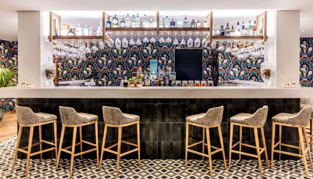 Interior view of a stylish bar featuring a polished marble countertop, high stools, and a decorative wall with colorful patterns, showcasing various bottles and glasses on display.