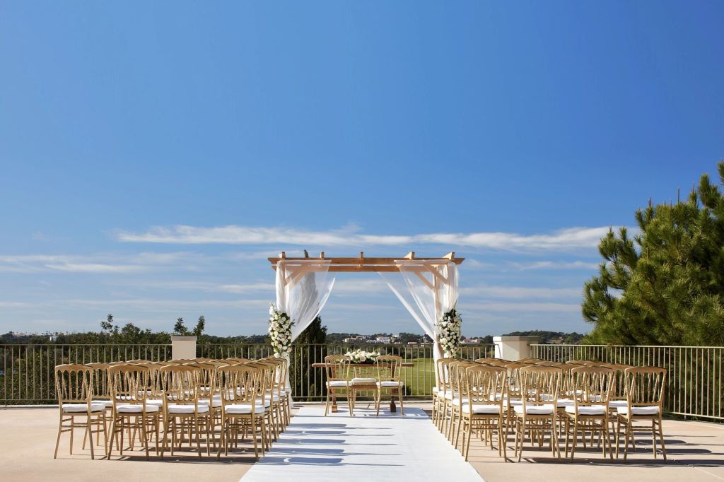 An outdoor wedding setup featuring a white aisle, decorated arch, and elegant chairs overlooking a scenic landscape under a clear blue sky.