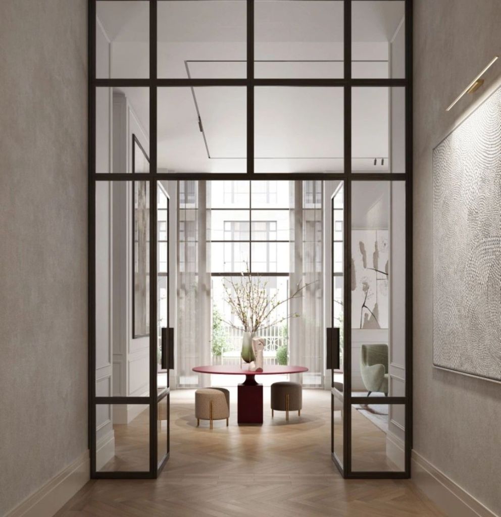 A modern entryway featuring tall glass doors, a stylish red table surrounded by elegant seating, and a decorative vase with flowers, showcasing a blend of luxury and contemporary design.