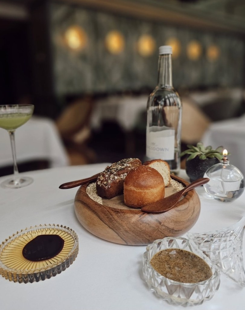 An elegant dining table setting showcasing a wooden bowl filled with different types of bread, accompanied by two dipping sauces. A bottle of water and a cocktail in a glass are also visible in the background, adding to the sophisticated atmosphere.