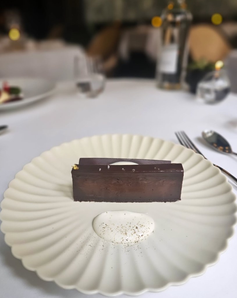 A beautifully plated chocolate dessert on a white scalloped plate, featuring a rectangular chocolate piece with a glossy finish and a dollop of cream beside it, sprinkled with a hint of spice.