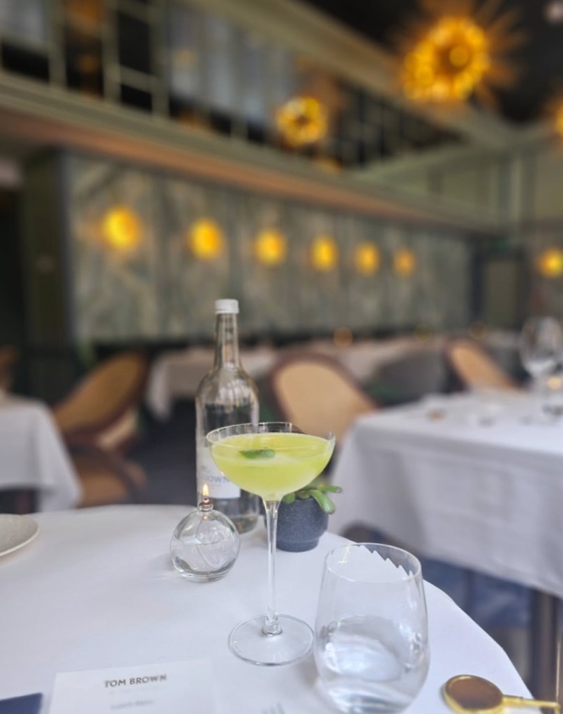A cocktail sits elegantly on a table at Tom Brown at The Capital, showcasing a vibrant yellow drink garnished with mint and accompanied by a stylish glass bottle of water, a candle, and a small succulent plant.
