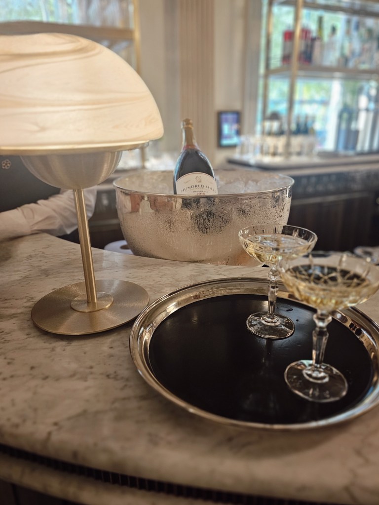 A stylish bar setup featuring a well-chilled bottle of Hundred Hills wine in a silver ice bucket, with two elegant champagne glasses on a silver tray, set against a marble surface and warm lighting.
