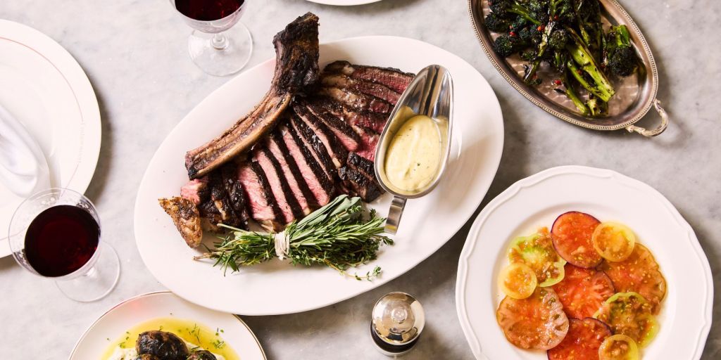 A beautifully plated meal featuring a large serving of grilled steak, sliced and arranged on a white plate, accompanied by a bowl of creamy sauce, fresh herbs, and sides of seasonal vegetables and heirloom tomatoes.