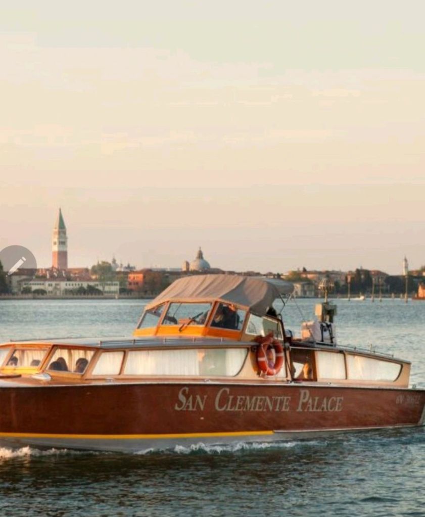 A wooden boat named 'San Clemente Palace' navigates the Venetian Lagoon, with historic buildings visible in the background against a soft sunset sky.