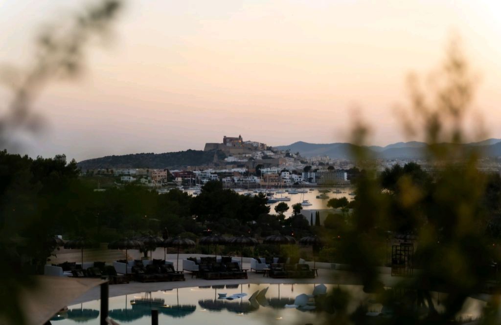 A scenic view of Ibiza's landscape at sunset, featuring a distant castle, boats in the harbor, and lush greenery in the foreground.