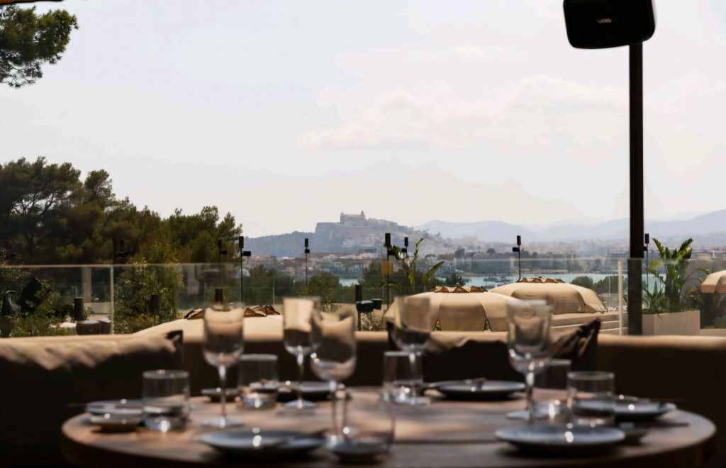 View of Ibiza from CLAP HOUSE with a dining table set in the foreground, showcasing glassware and plates against a backdrop of greenery and distant hills.