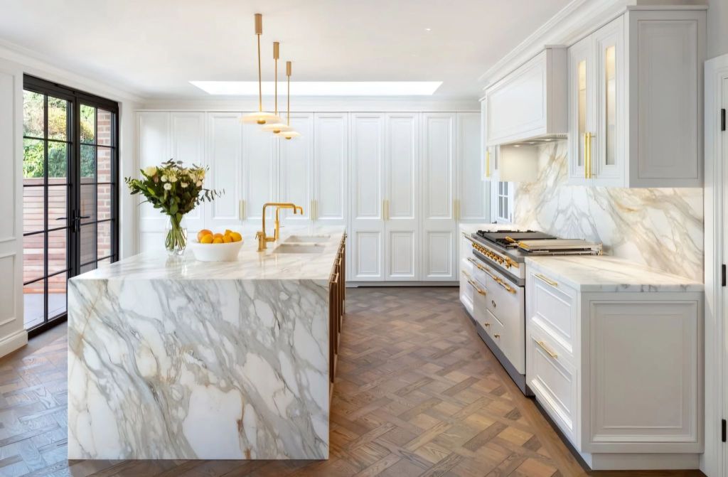 A modern kitchen featuring a large marble island, white cabinetry, and a Lacanche range, highlighted by elegant gold fixtures and a bouquet of flowers.