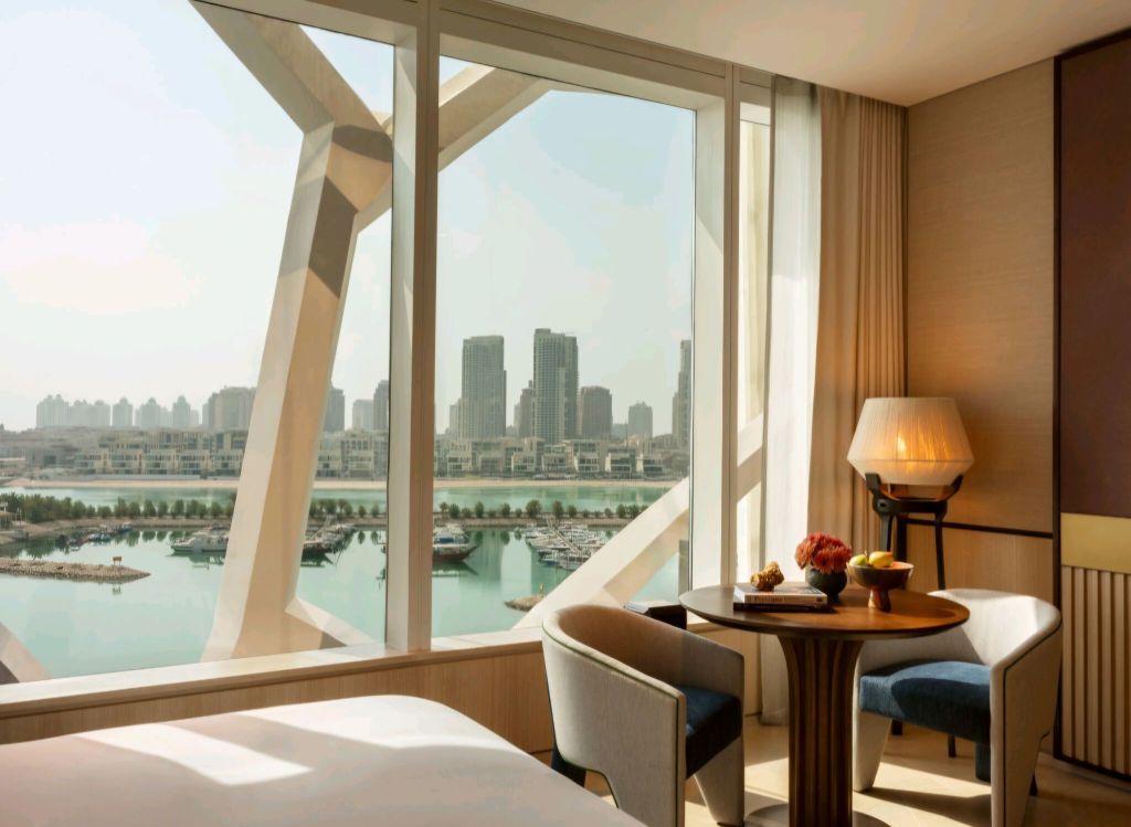Interior view of a luxurious hotel room featuring a seating area with a round table, decorative fruits, and a lamp, alongside large windows showcasing a scenic view of the marina and city skyline.