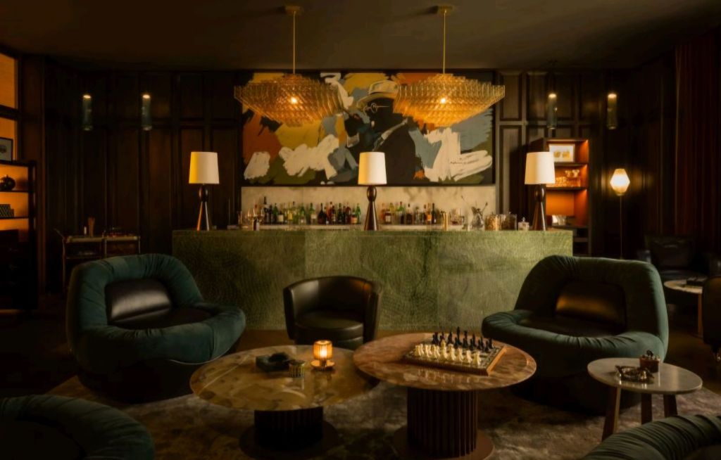 Interior view of a stylish bar area featuring plush seating, a green marble bar topped with a chandelier, and artistic decor on the walls.