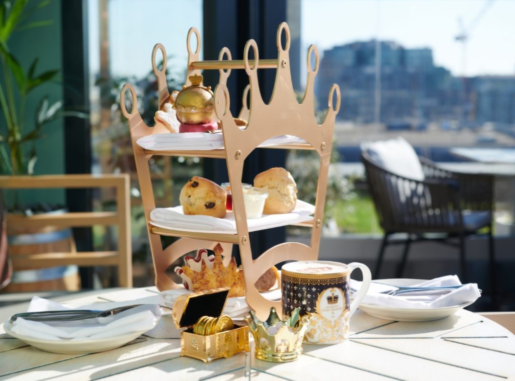 A beautifully presented afternoon tea setup on a table, featuring a three-tier stand adorned with scones and elegant decorations, alongside a royal-themed teacup and a golden crown.
