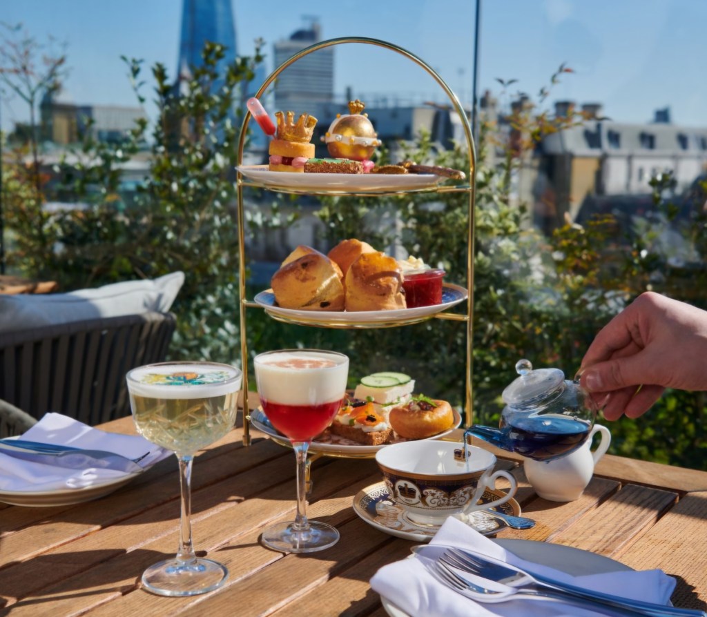 A beautifully presented afternoon tea set, featuring a tiered stand with various gourmet savories and pastries, including scones and decorative desserts, alongside elegantly crafted cocktails and a pot of tea, displayed on a wooden table with a cityscape in the background.