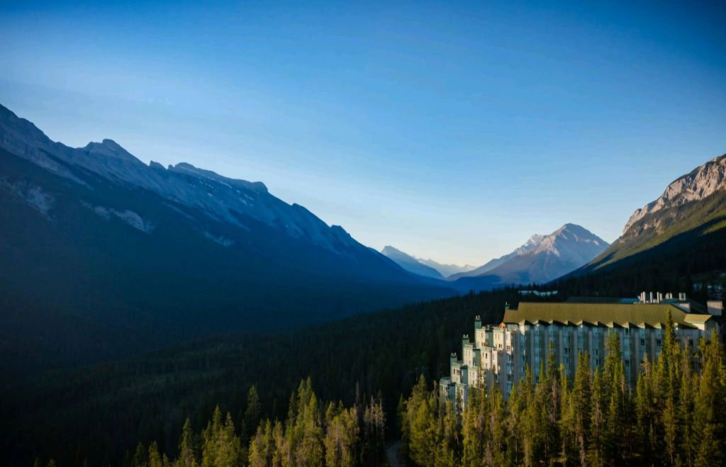 A panoramic view of The Rimrock Resort Hotel nestled in a forested valley, surrounded by majestic mountains and a clear blue sky.