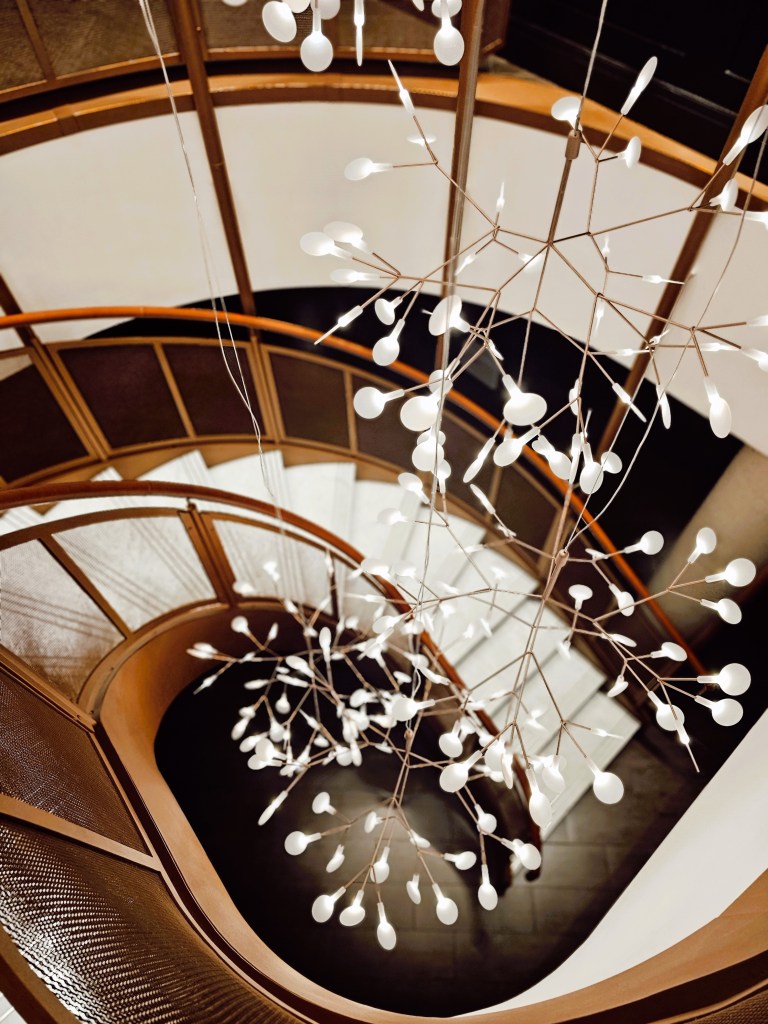 A view from above of a spiral staircase featuring a modern chandelier with white light fixtures, highlighting the elegant design and warm ambiance of a hotel interior.